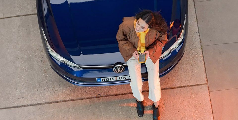 Junge Frau mit Handy in der Hand, lehnt sich an dunkelblauen Volkswagen Neuwagen